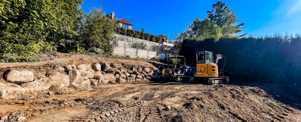 Deux mini-excavatrices sur un terrain résidentiel près d’un mur de soutènement en pierre.