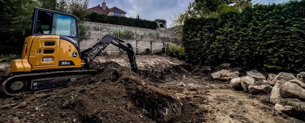 Mini-excavatrice nivelant un terrain résidentiel près d’un mur de soutènement.