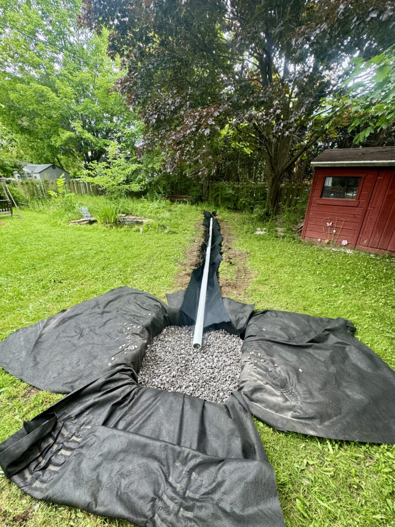 Installation d’un drain de surface avec géotextile, pierre nette et tuyau de drainage dans une cour résidentielle.