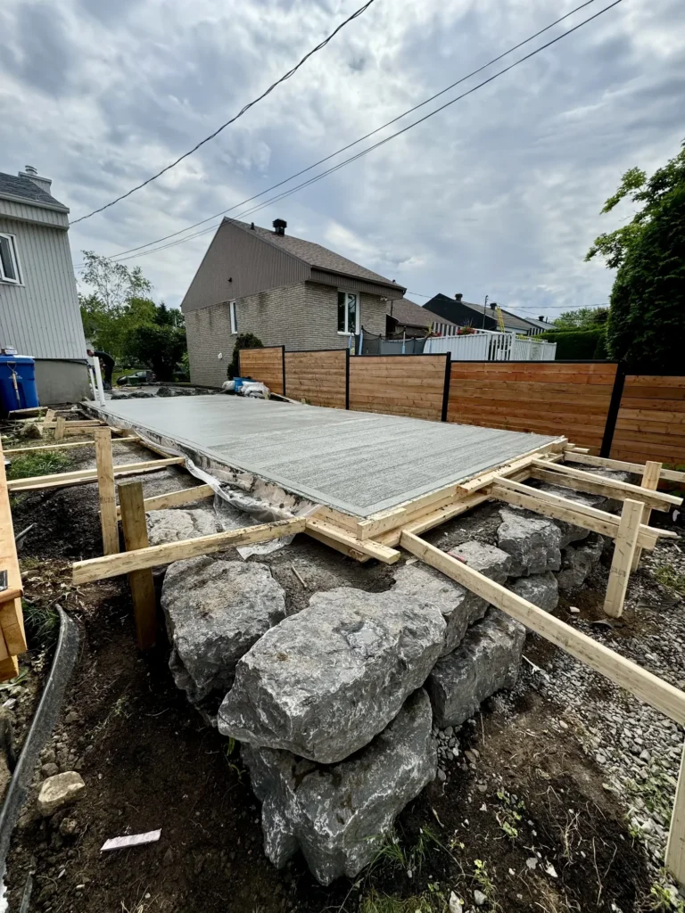 Nouvelle dalle de béton coulée dans une cour arrière avec coffrage en bois, muret de pierres et clôture en bois