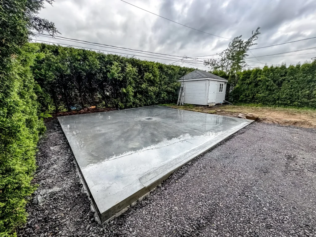 Dalle de béton lissée fraîchement coulée dans une cour arrière avec cabanon gris et haie de cèdres