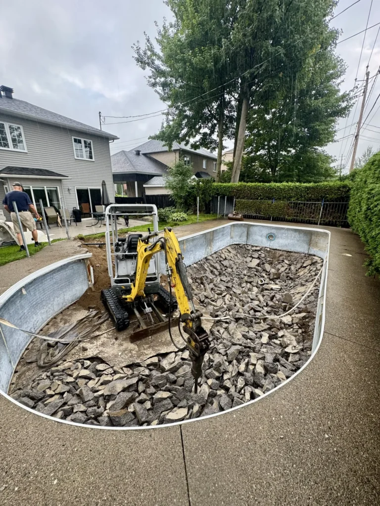 Mini-excavatrice jaune démolissant une piscine creusée remplie de gravats dans une cour arrière sous la pluie