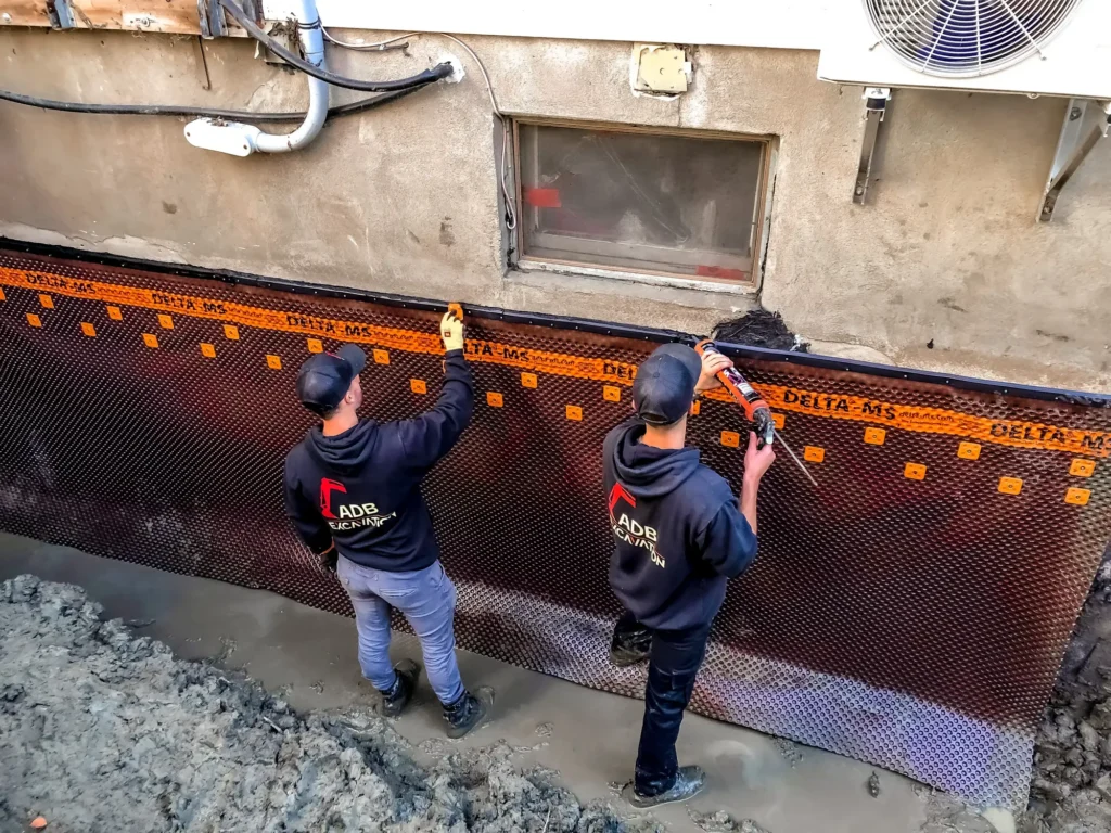 Deux ouvriers ADB Excavation installent une membrane de drainage Delta-MS sur un mur de fondation