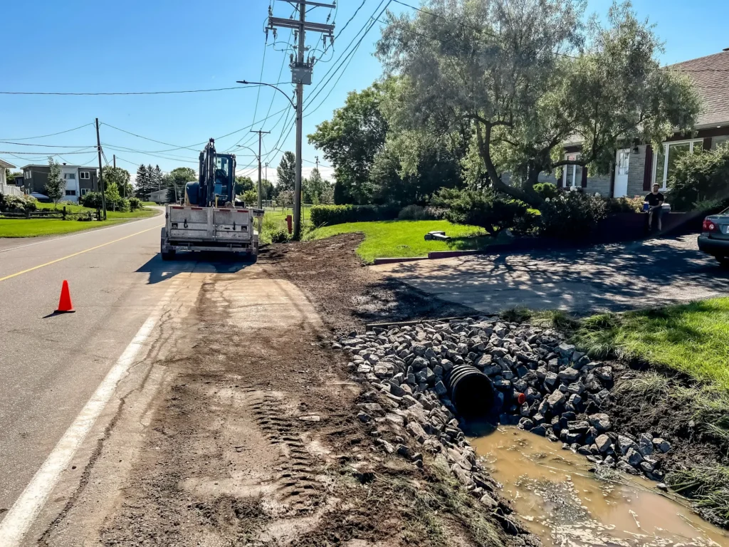 Chantier d'installation de ponceau avec enrochement et drainage en bordure de route résidentielle, camion et excavatrice