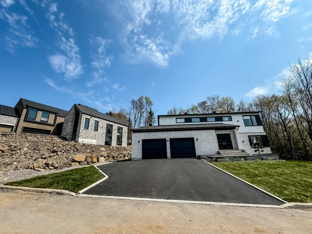 Maison moderne de luxe avec façade blanche, garage double noir et longue allée pavée sous ciel bleu