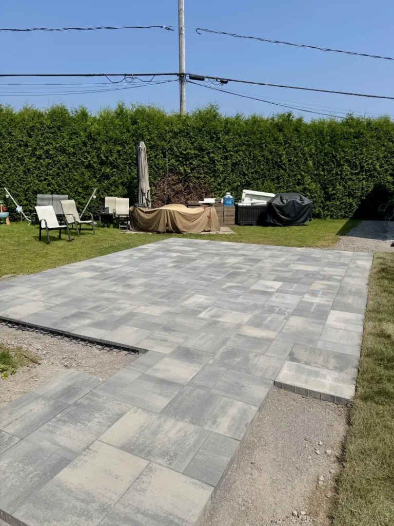 Patio en pavés gris récemment installé dans une cour arrière avec haie de cèdres et mobilier de jardin