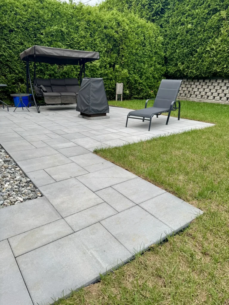 Terrasse en dalles de béton gris avec balancelle, chaise longue et haie de thuyas dans jardin paysager